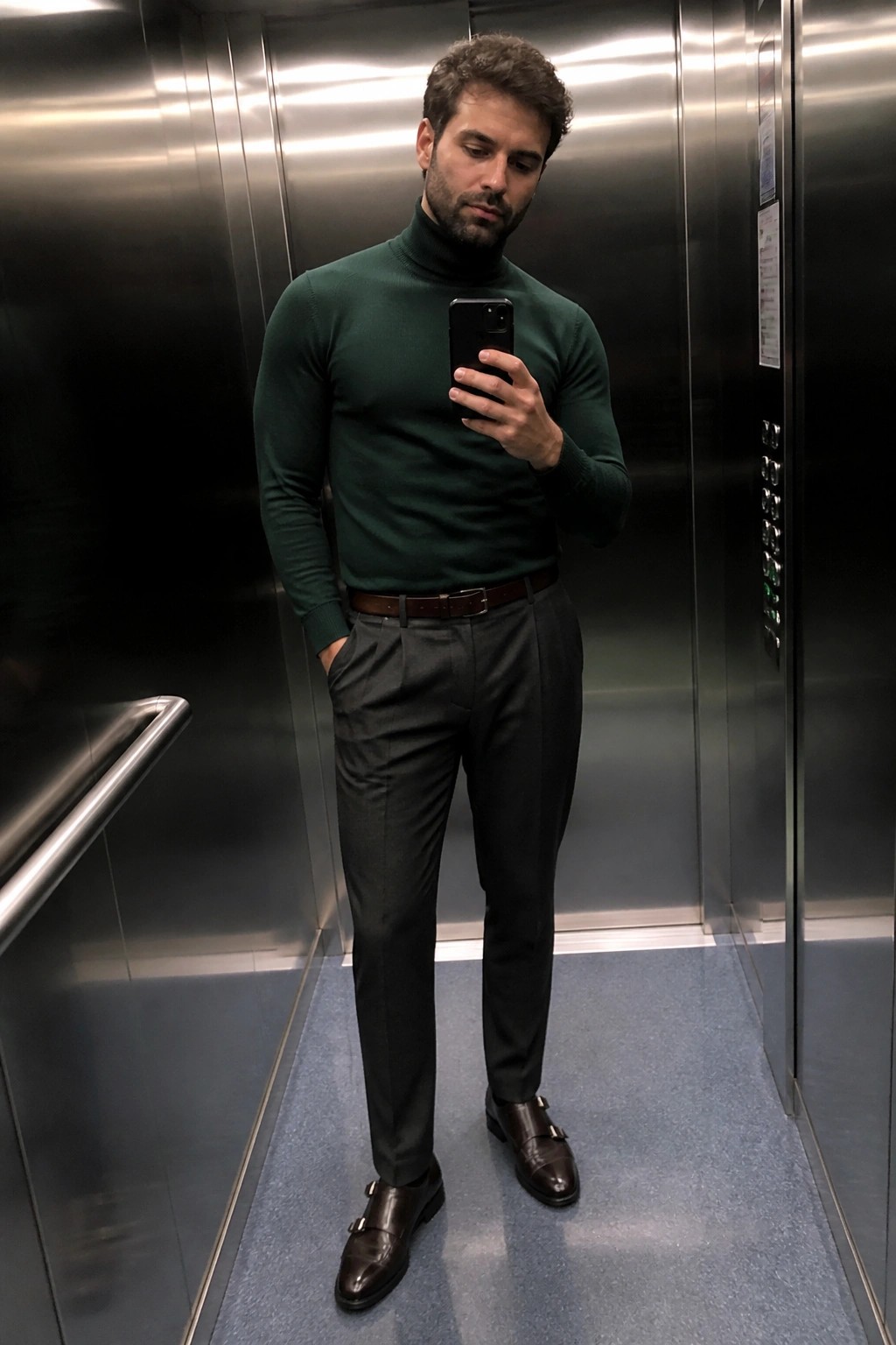 Man in green turtleneck and gray trousers taking selfie in elevator.