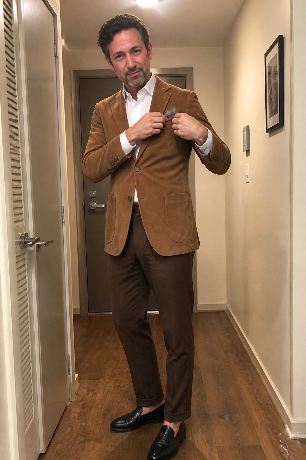 Man in brown suede blazer and trousers adjusting lapel in hallway