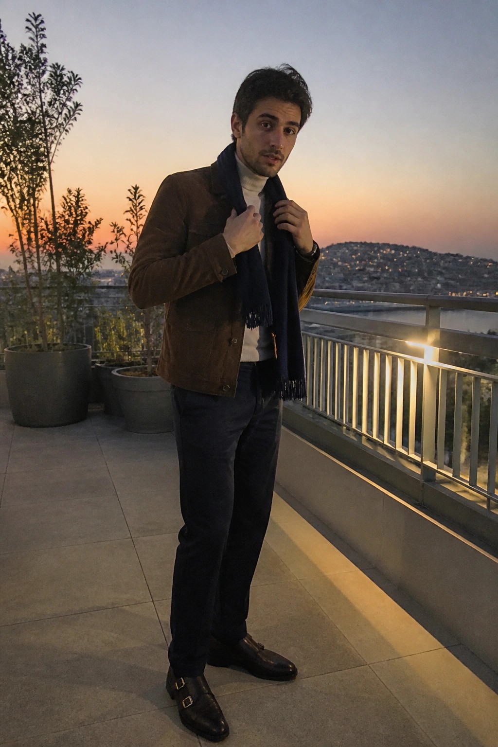 Man in brown suede jacket and blue scarf adjusts clothing on sunset balcony