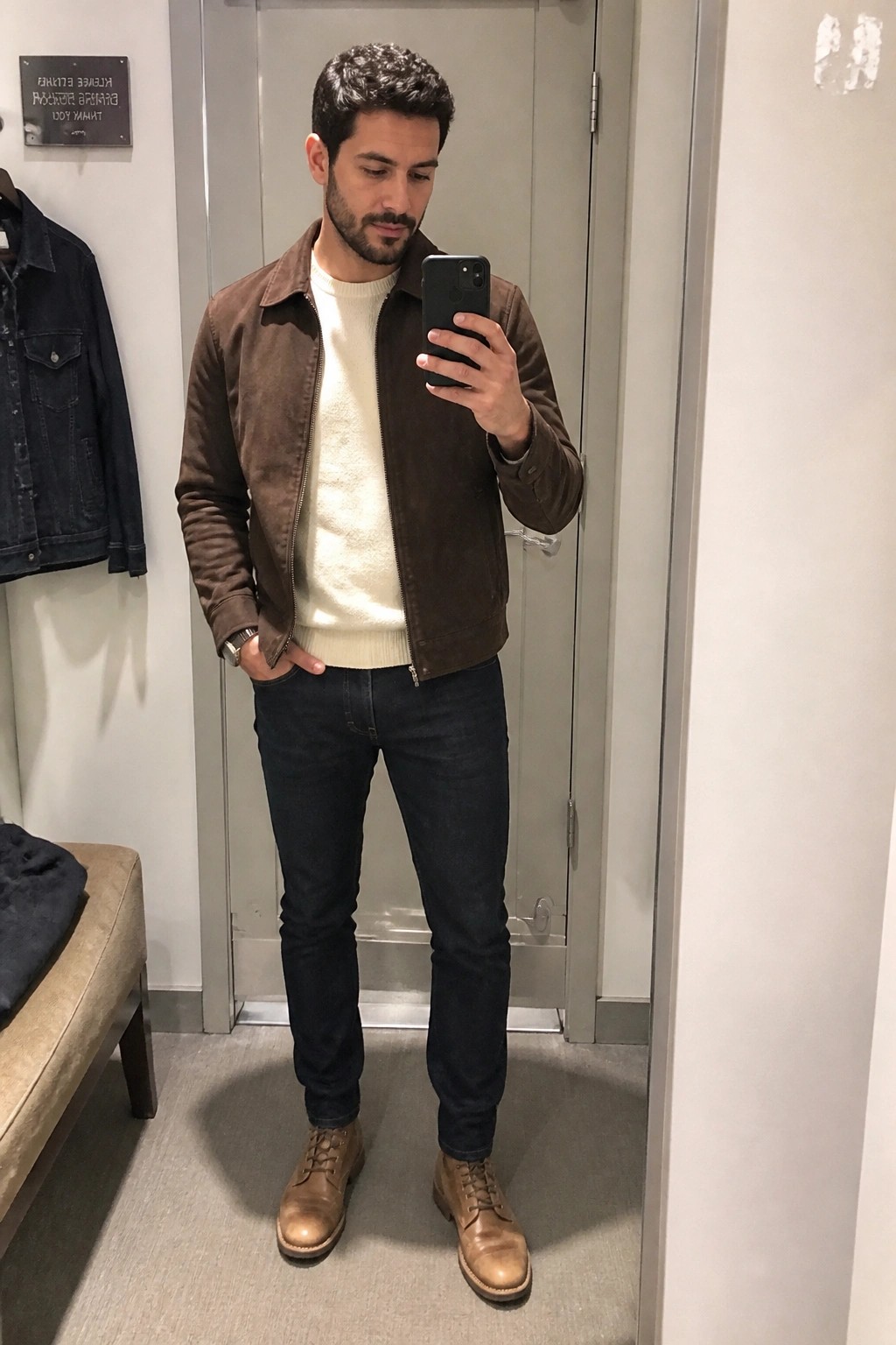 Man in brown suede jacket over cream sweater with dark jeans and brown leather boots taking a mirror selfie.