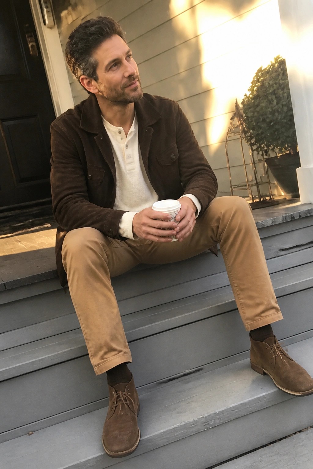 Man in brown jacket and khaki pants sits on gray porch steps holding coffee