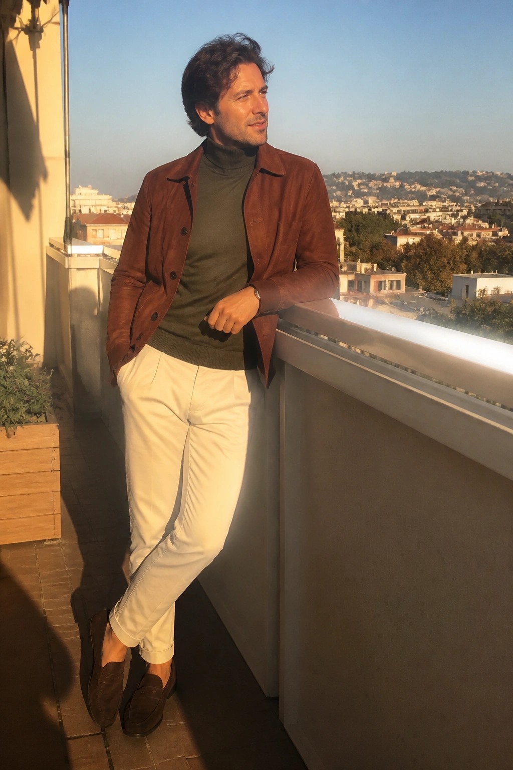 Man in brown suede jacket, green turtleneck, and white pants on sunny balcony.