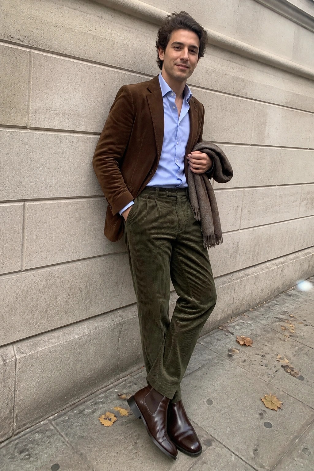 Man in brown velvet blazer, blue shirt, green corduroy pants by stone wall.