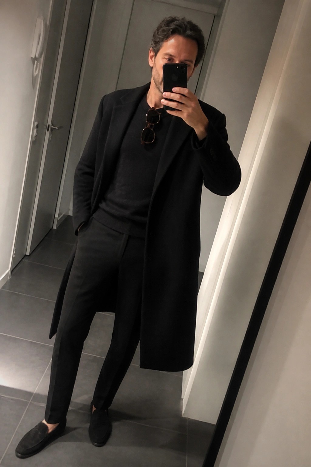 Man in black overcoat and sweater taking mirror selfie in hallway