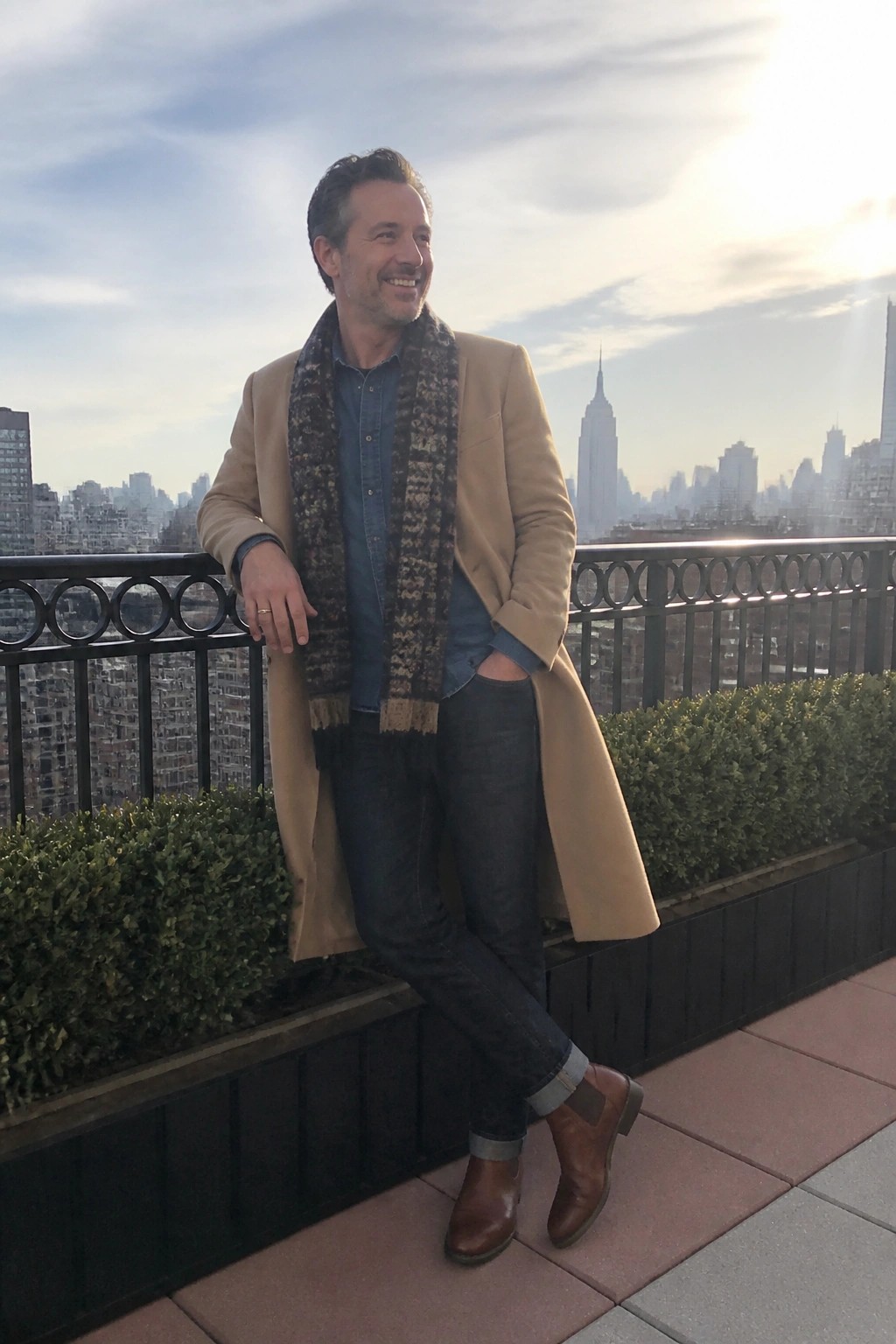 Smiling man in tan coat and patterned scarf on rooftop overlooking city skyline