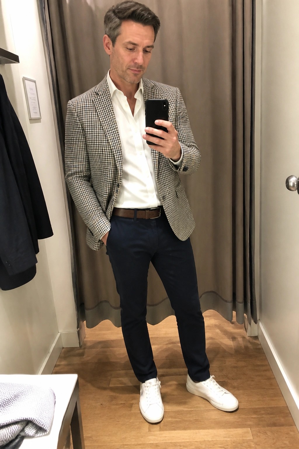 Man in houndstooth blazer, white shirt, navy pants, and white sneakers holds phone.