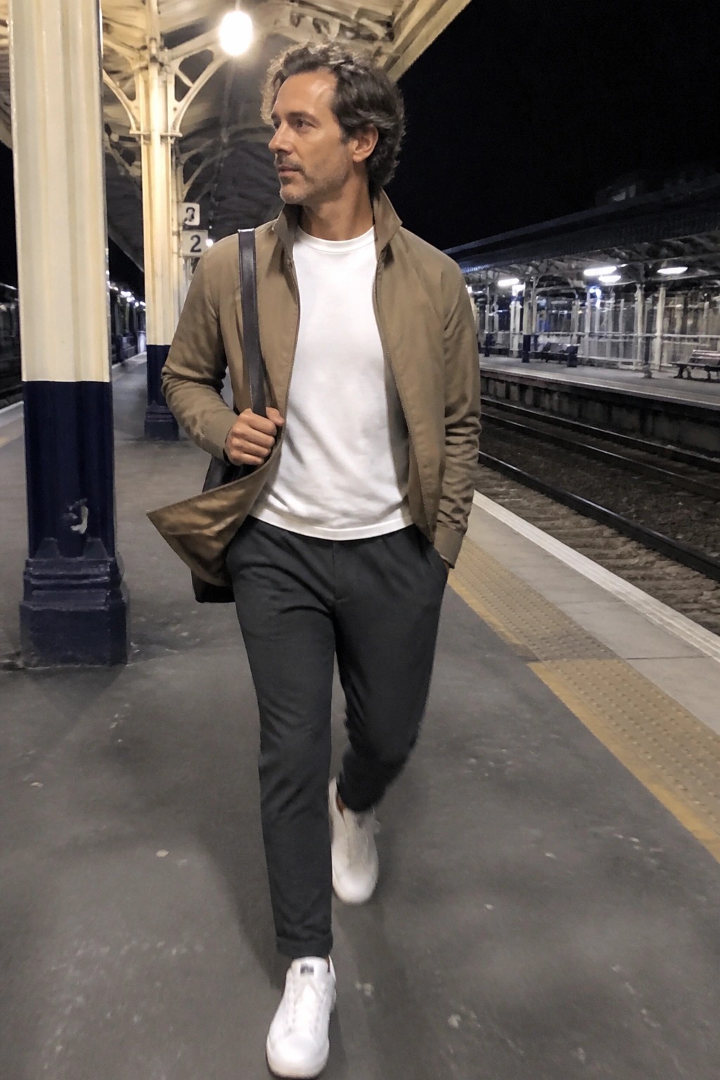 Man in open olive jacket over white tee walks on nighttime train platform