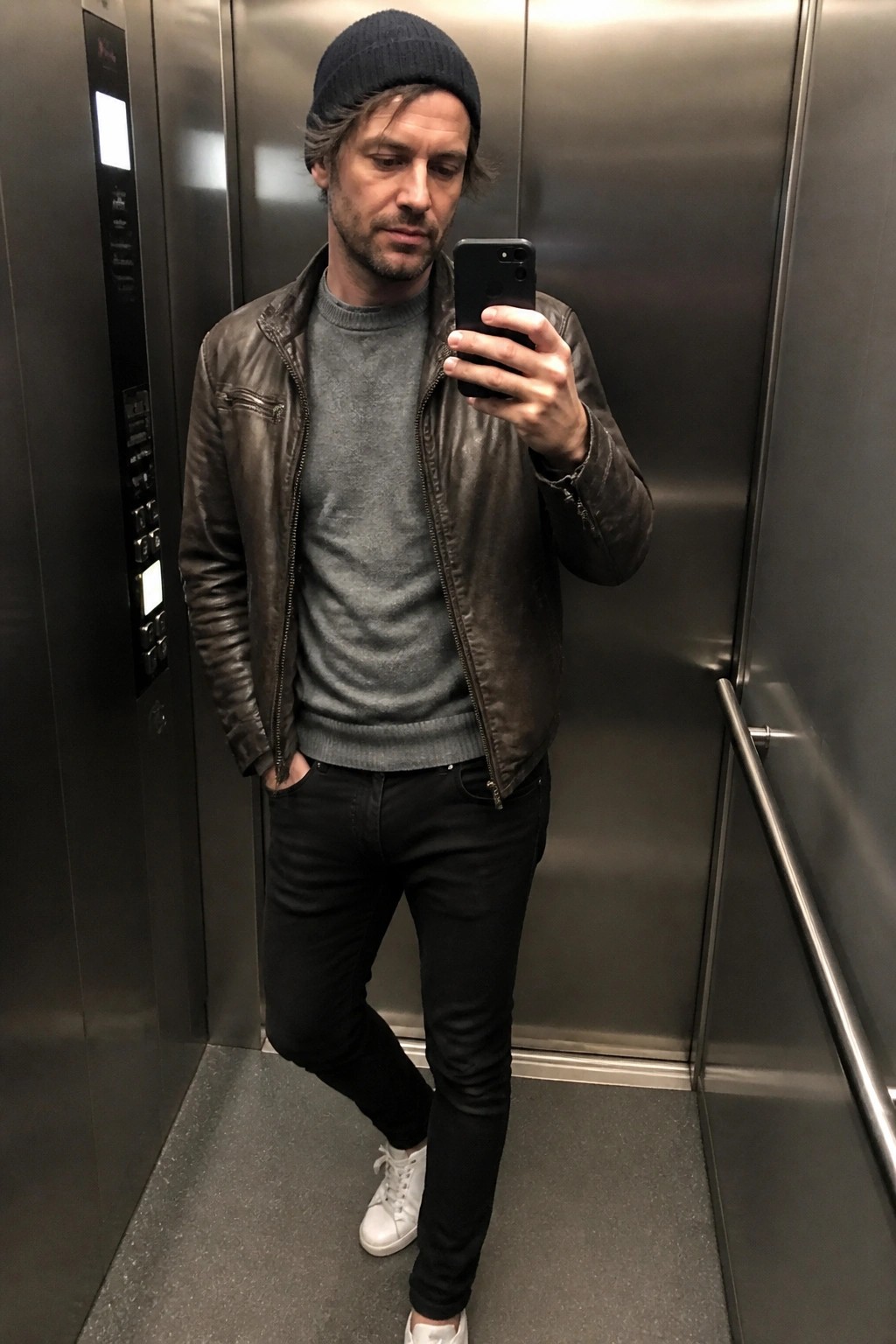 Man in brown leather jacket, beanie, and jeans taking elevator selfie.