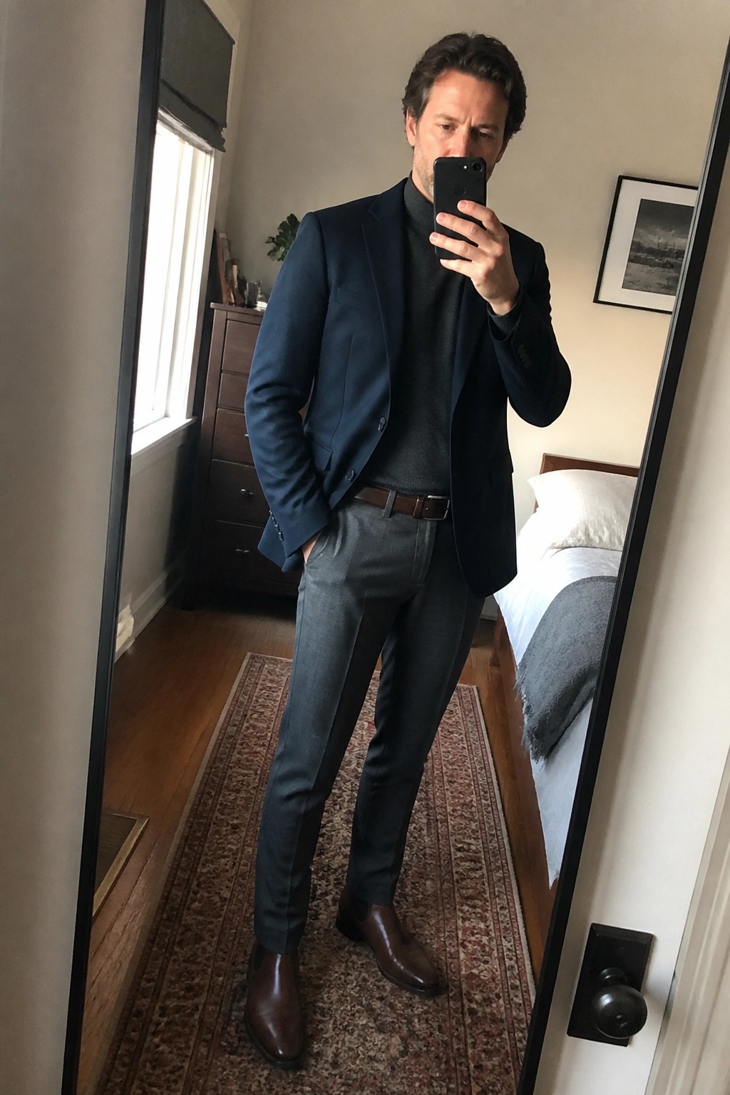 Man in navy blazer over turtleneck with gray pants taking mirror selfie in bedroom