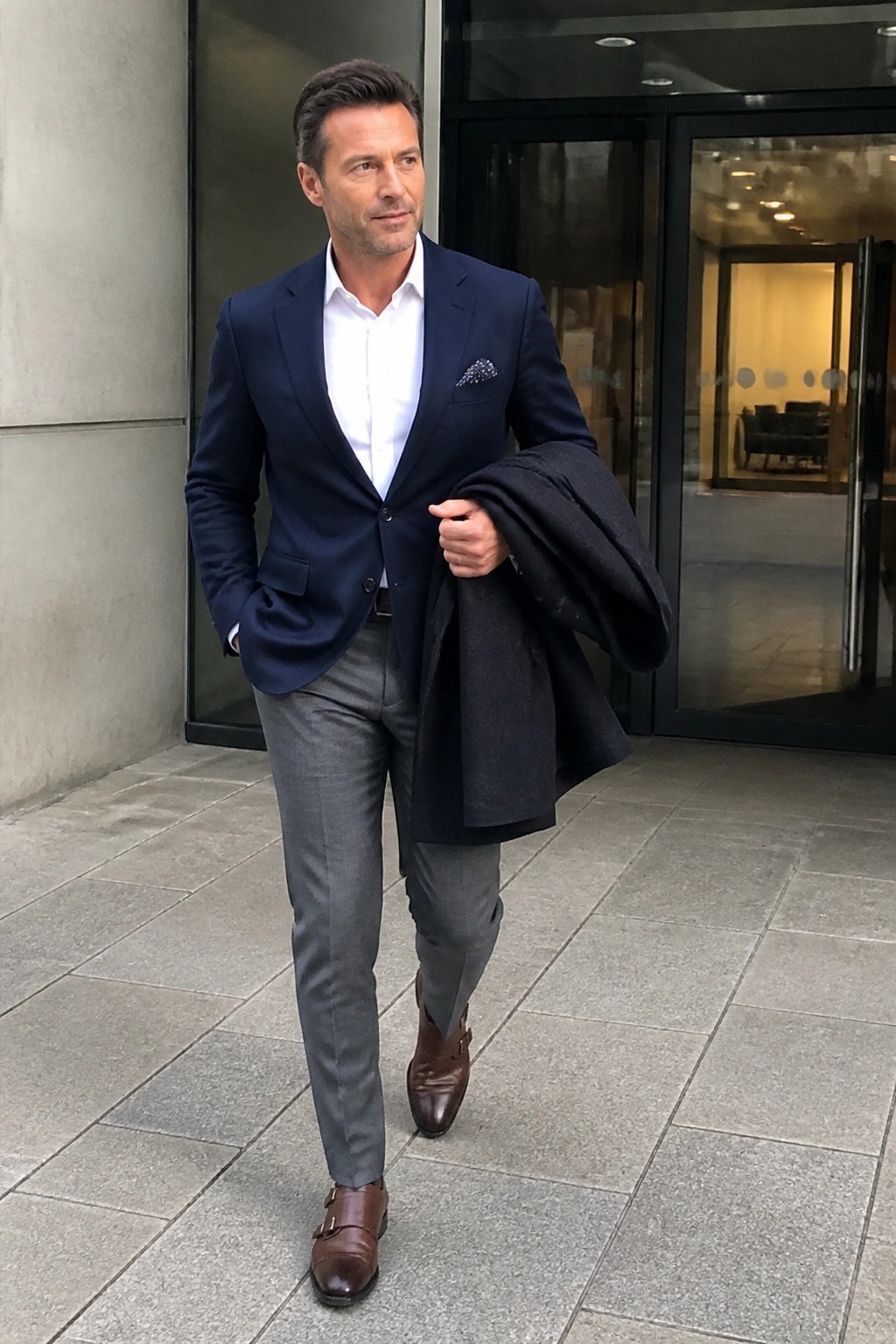 Man in navy blazer holding dark coat outside glass building entrance