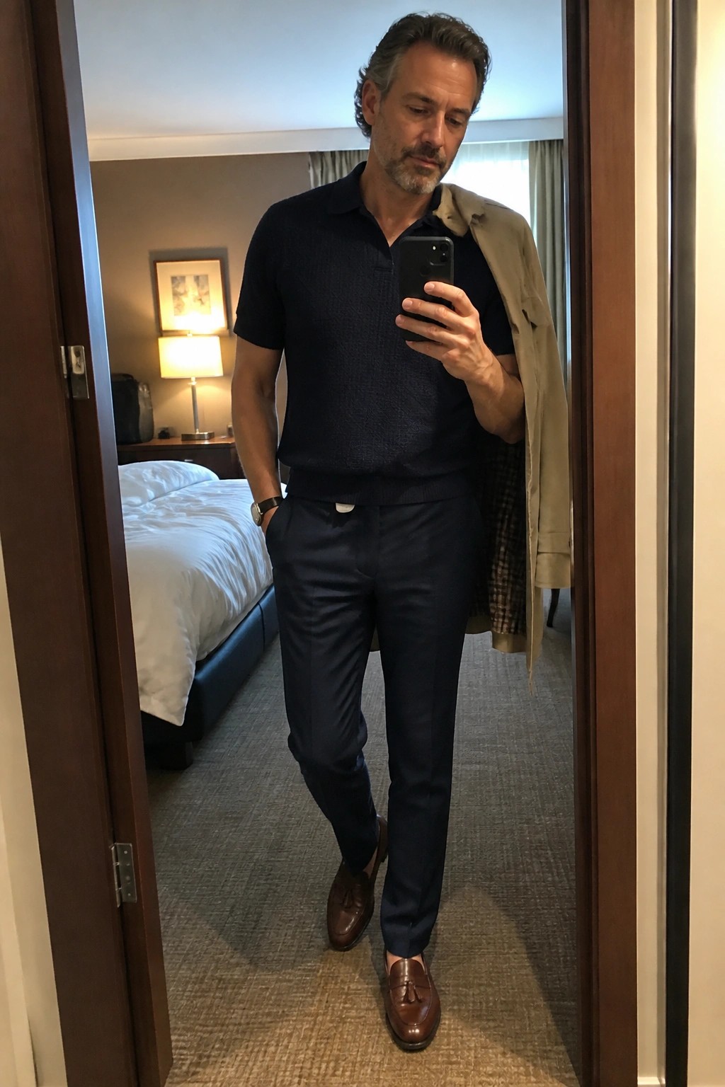 Man in navy sweater and trousers taking mirror selfie with beige jacket over shoulder