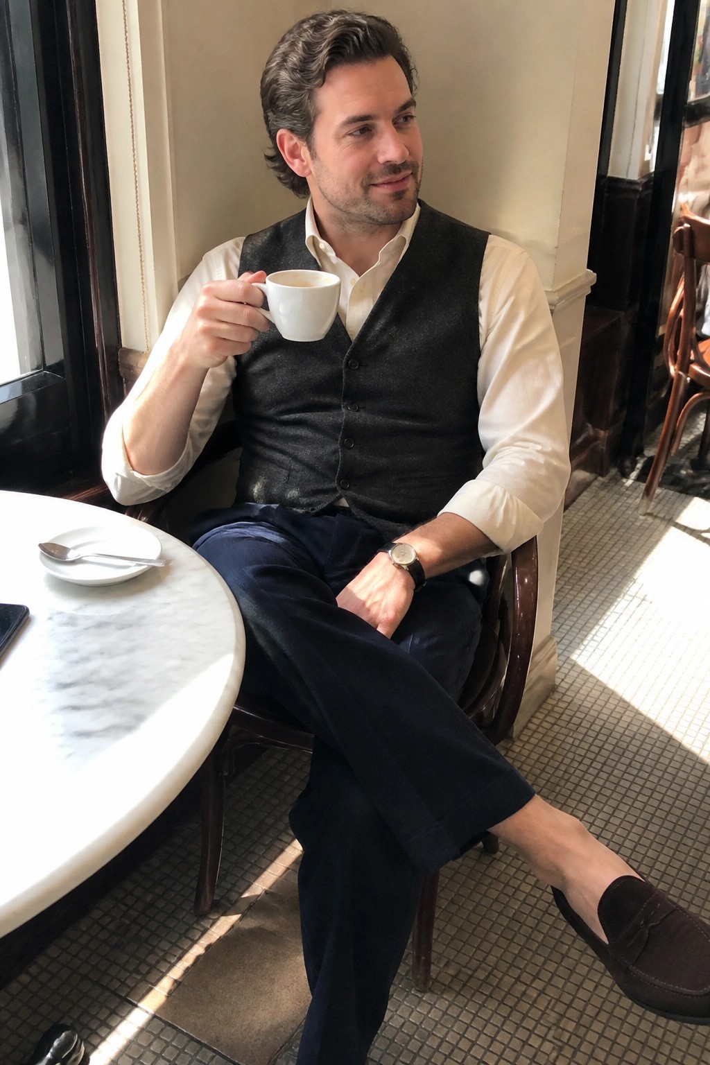 Man seated in a cafe wearing a white button down shirt under a dark gray wool vest, navy trousers, brown suede loafers, and a wristwatch while holding a white coffee cup.