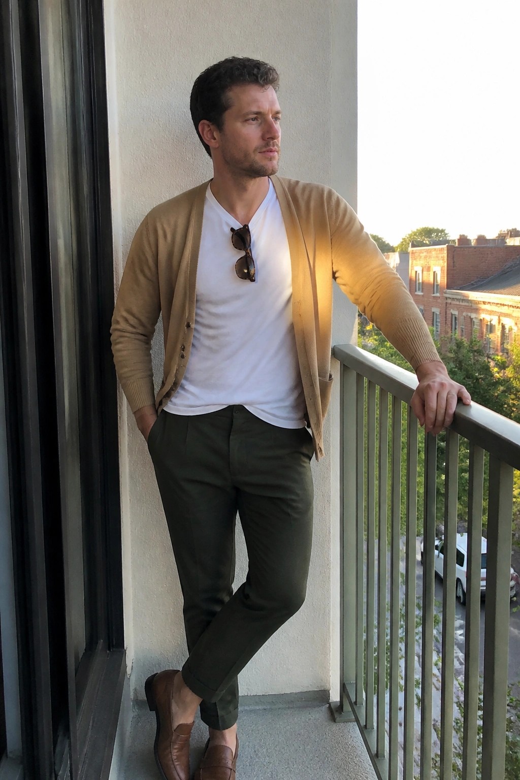 Man in tan cardigan over white v-neck tee, olive trousers, and brown loafers standing on a balcony with sunglasses tucked into the shirt.