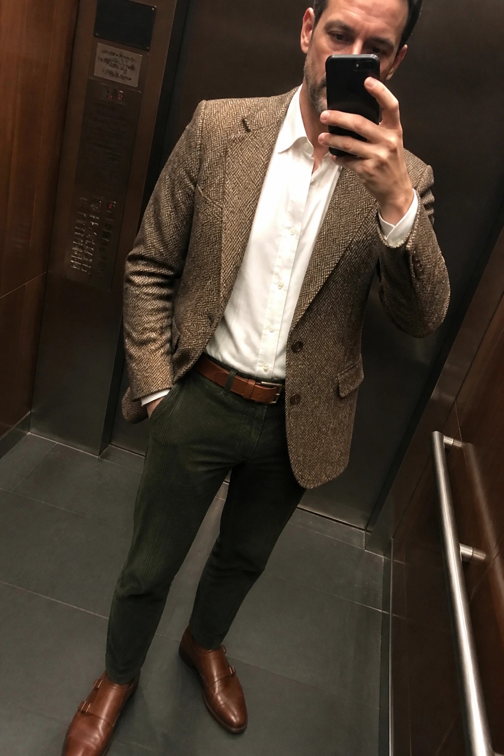 Man in brown herringbone blazer, white shirt, green pants taking elevator selfie