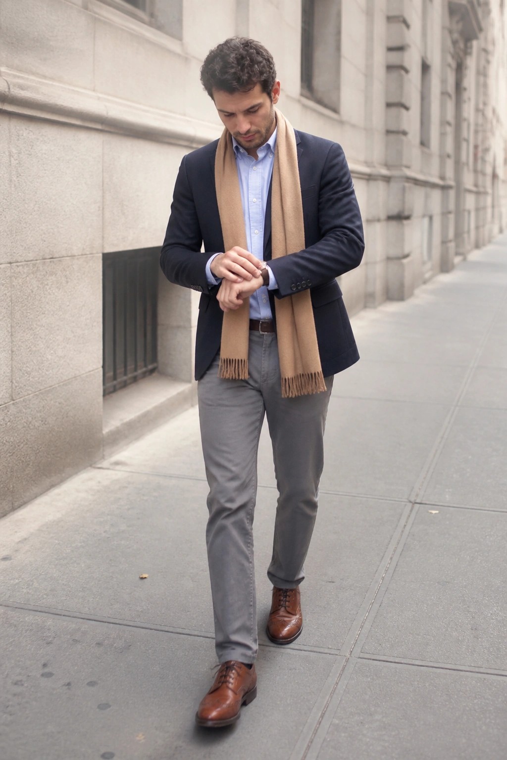 Man in navy blazer and beige scarf checks watch on city sidewalk