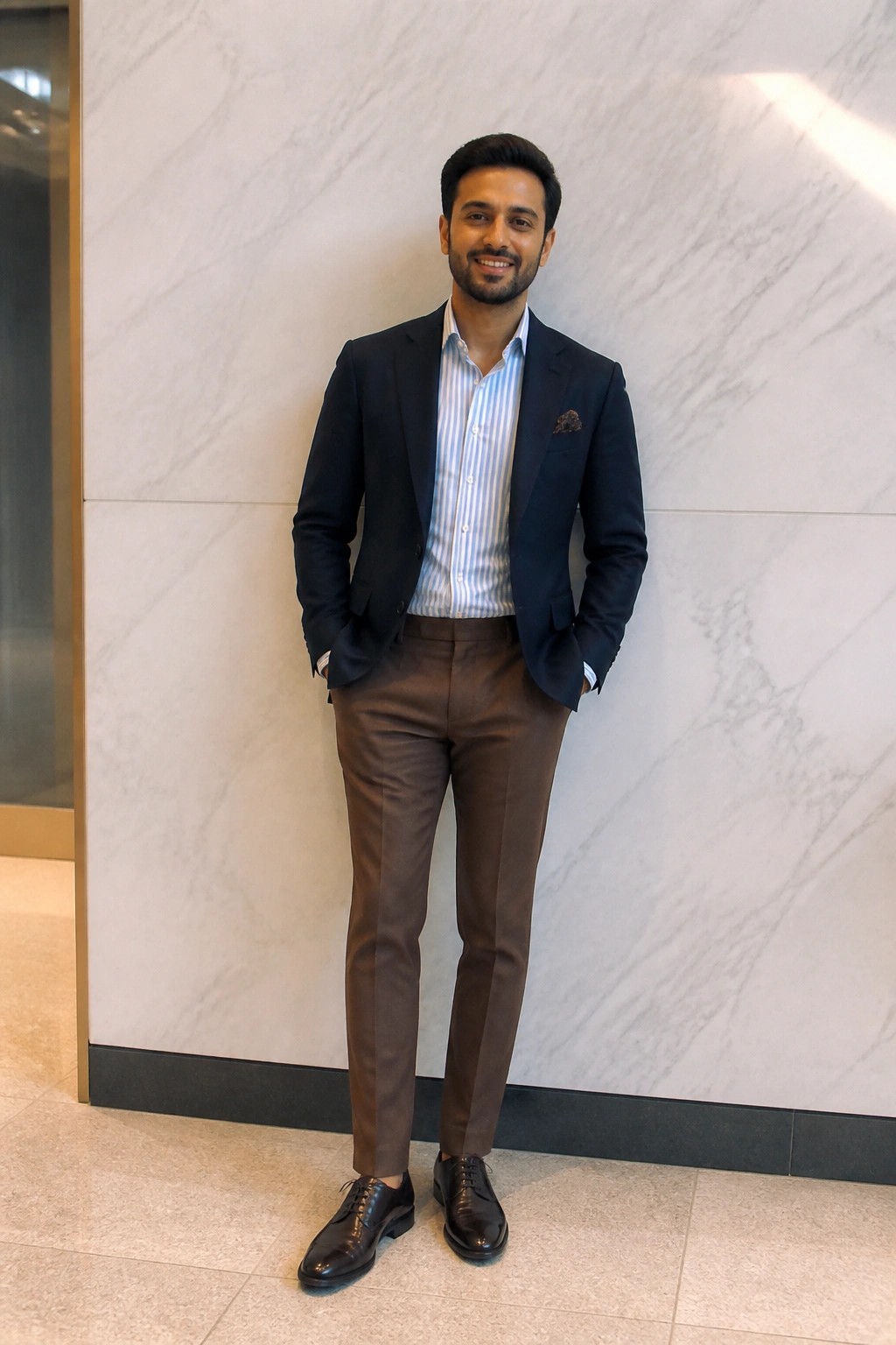 Man in navy blazer, striped shirt, brown pants posing against marble wall