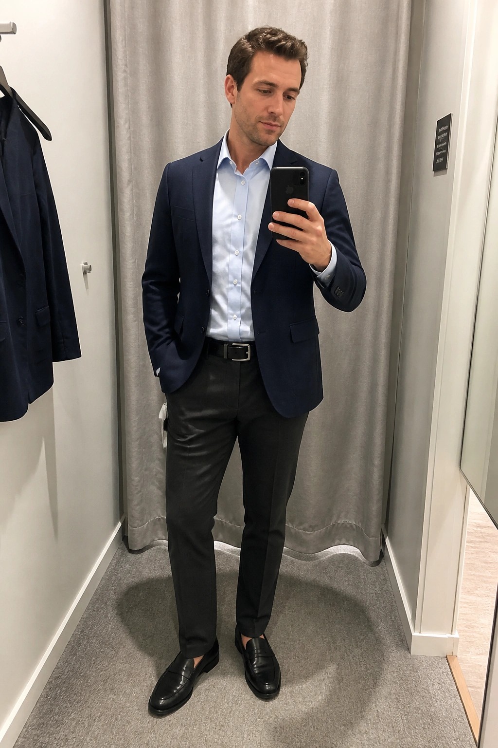 Man in navy blazer and light blue shirt takes mirror selfie in fitting room.