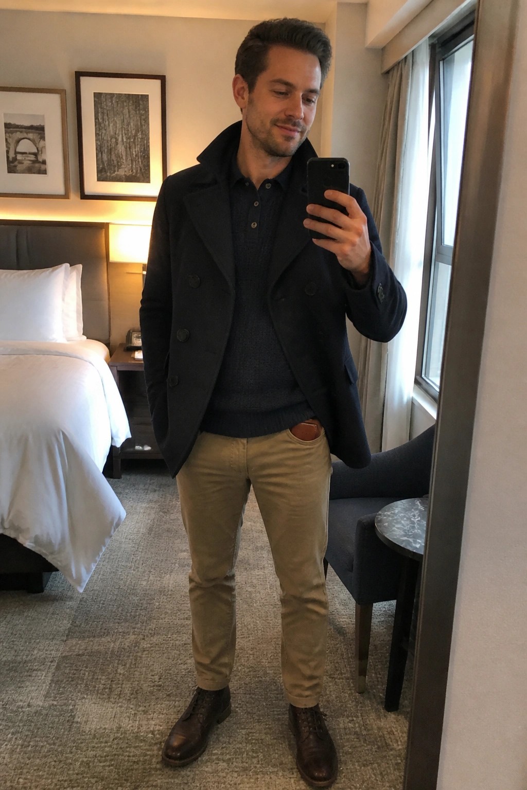 Man wearing a navy double-breasted peacoat over a dark blue knit sweater with khaki pants and brown leather shoes while holding a phone in a hotel room setting.