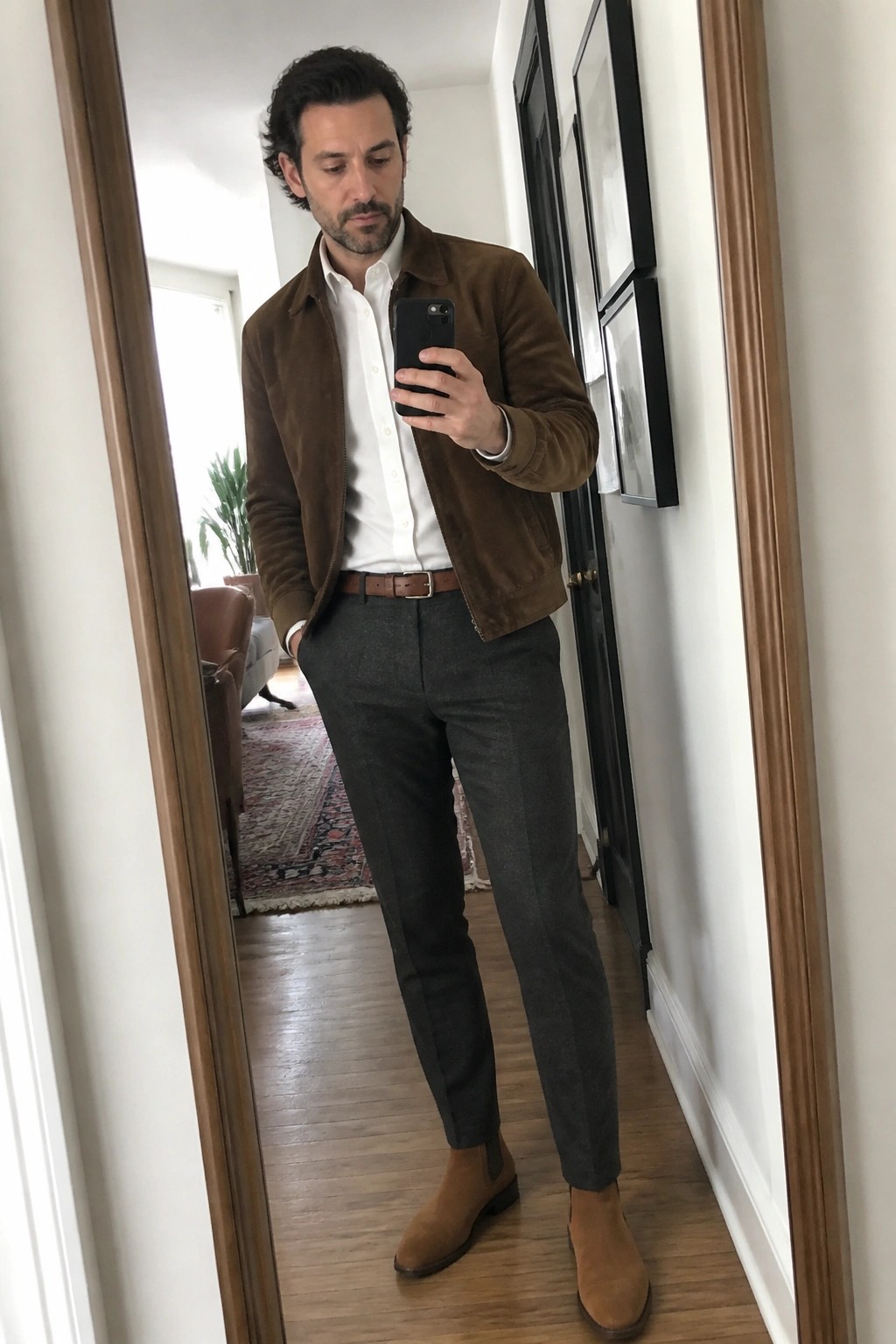 Man in brown suede jacket, white shirt, gray pants, and boots taking mirror selfie