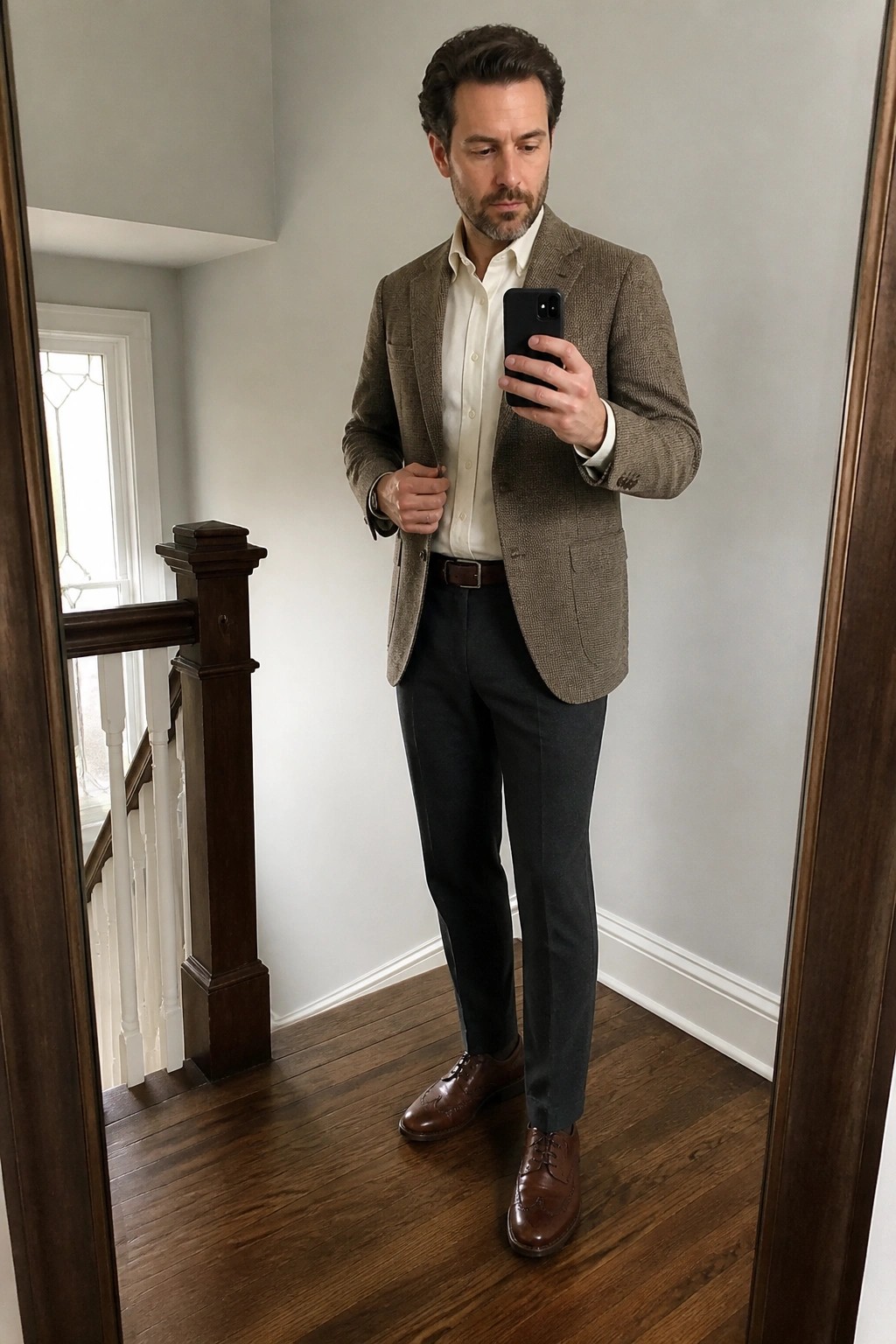 Man in brown tweed blazer, white shirt, and dark pants holding smartphone in mirror