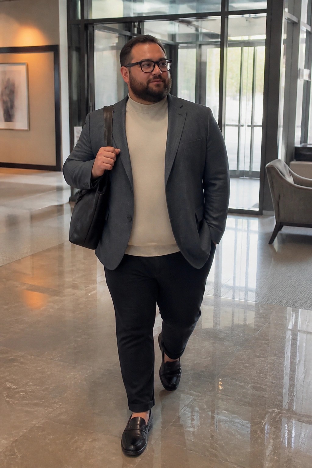 Bearded man in gray blazer and beige turtleneck walking in modern marble lobby with bag