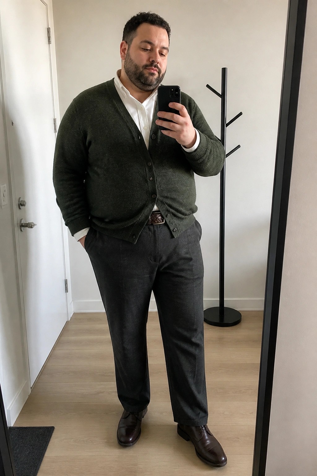 Man in green cardigan over white shirt taking mirror selfie in gray trousers.