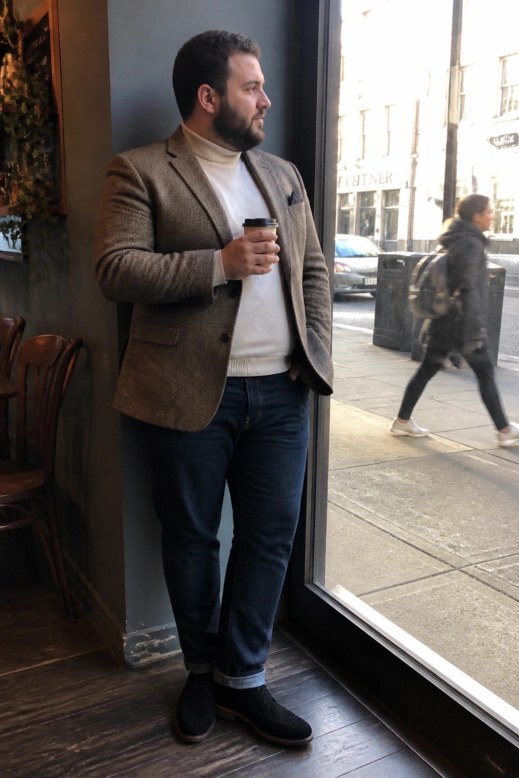 Bearded man in brown herringbone blazer and jeans holds coffee by cafe window.