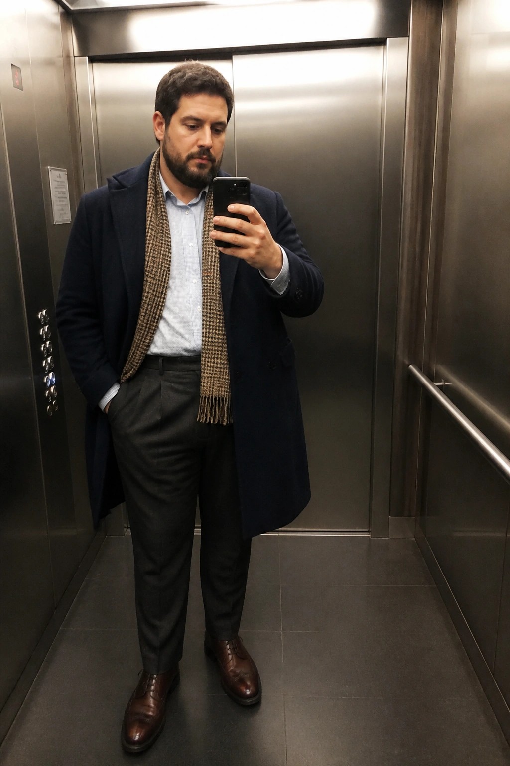 Man in navy coat and houndstooth scarf taking phone selfie in elevator