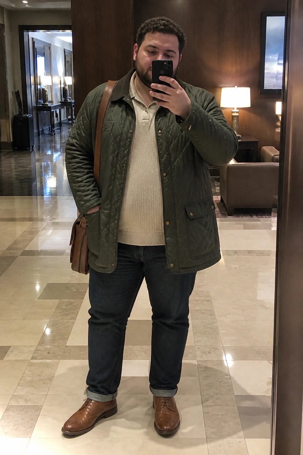 Man in green quilted jacket taking mirror selfie with leather bag in lobby