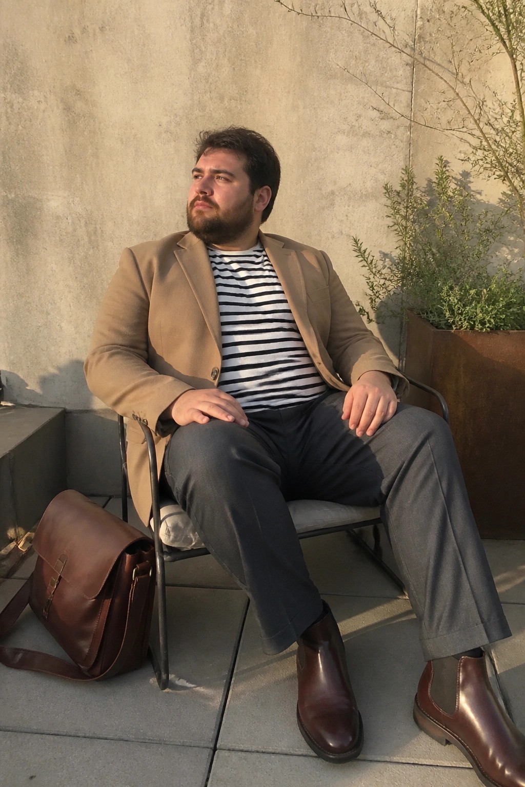 Bearded man in tan blazer over striped shirt sits outdoors beside brown leather bag