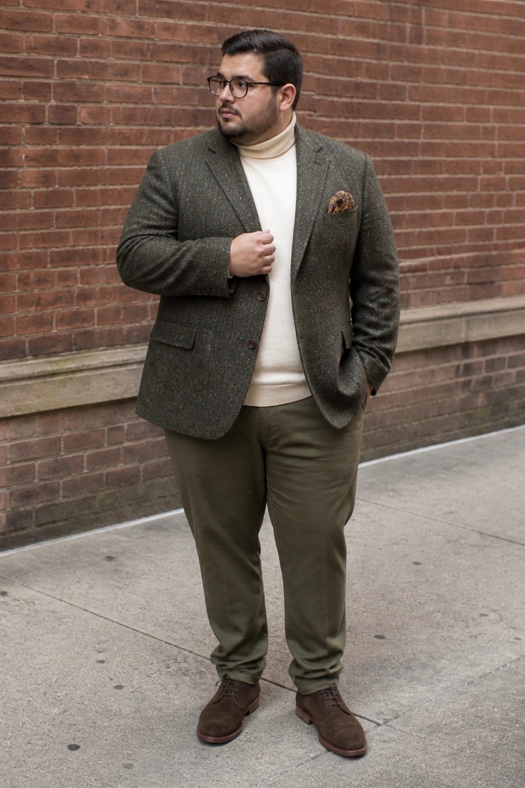 Man in green tweed blazer, cream turtleneck, and olive pants by brick wall.
