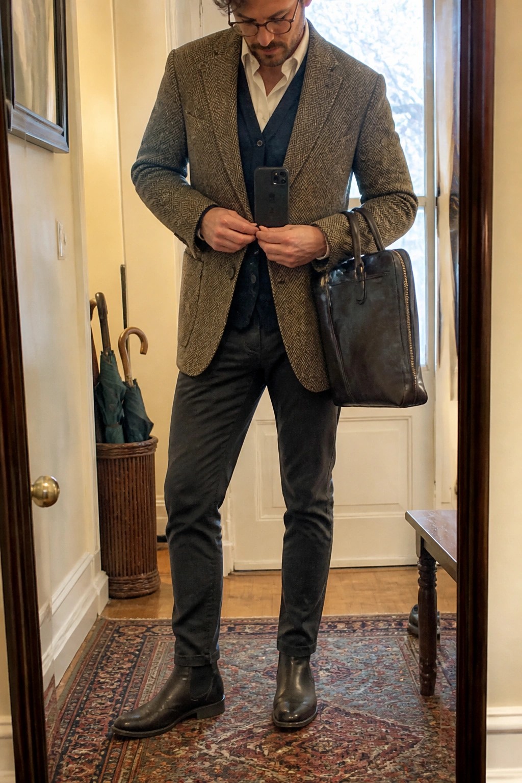 Man wearing a brown herringbone tweed blazer over a blue vest and white shirt with dark pants and black leather Chelsea boots while holding a dark leather bag.