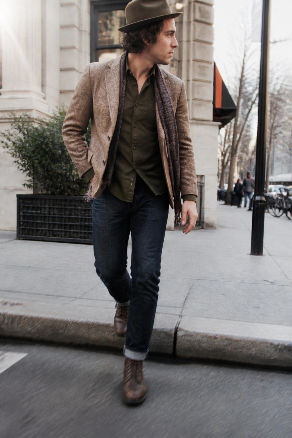 Man wearing a brown herringbone tweed jacket over an olive button down shirt with a dark patterned scarf, dark blue jeans cuffed at the ankle, brown leather boots, and a matching brown hat while walking on a city sidewalk.
