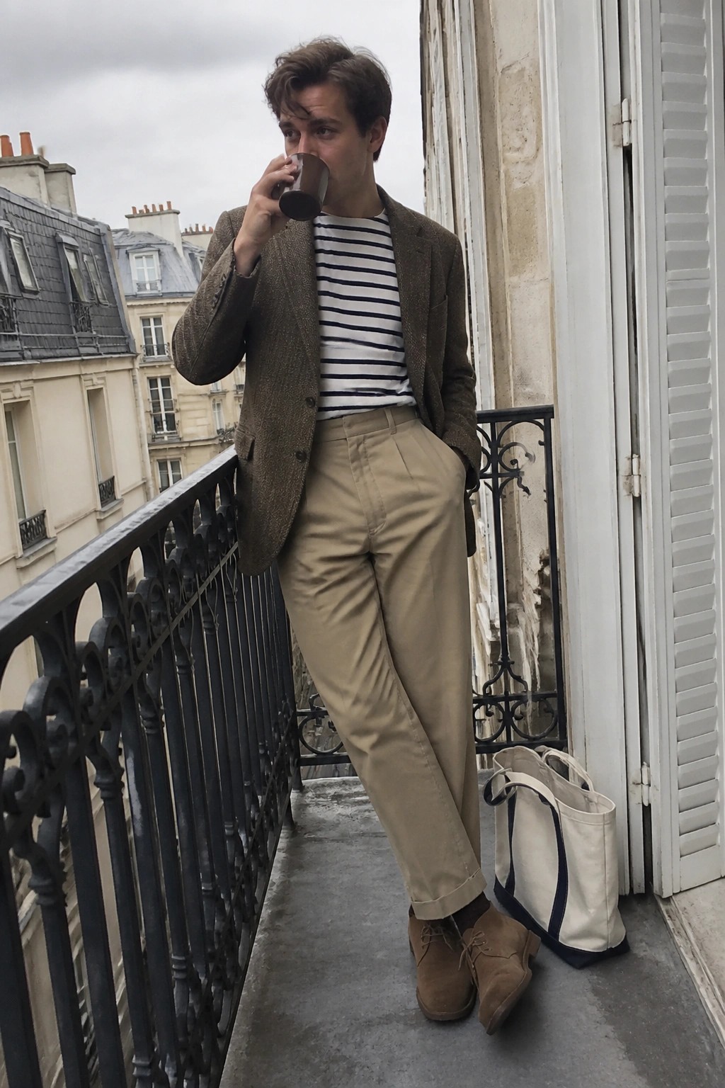 Man in brown tweed blazer and striped shirt drinks coffee on Paris balcony with tote bag.