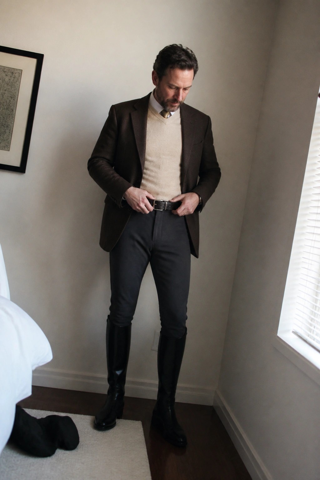 Man in brown blazer and beige sweater adjusts belt with black riding boots
