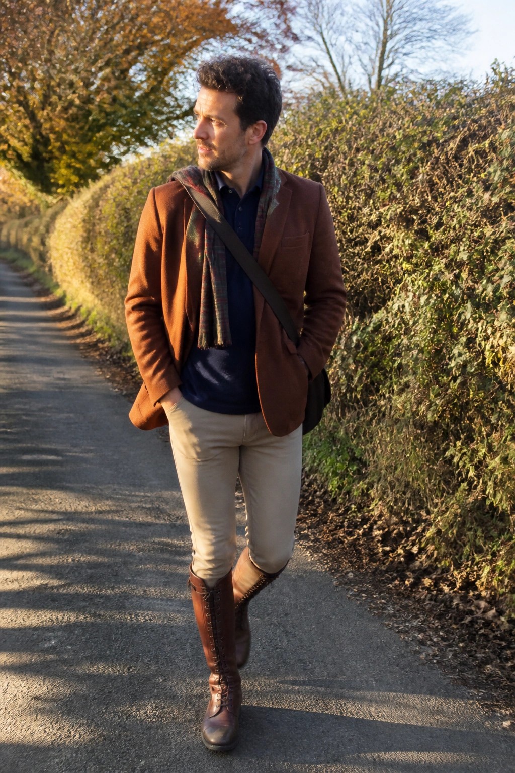 Man in brown blazer, plaid scarf, beige pants, tall boots on autumn path