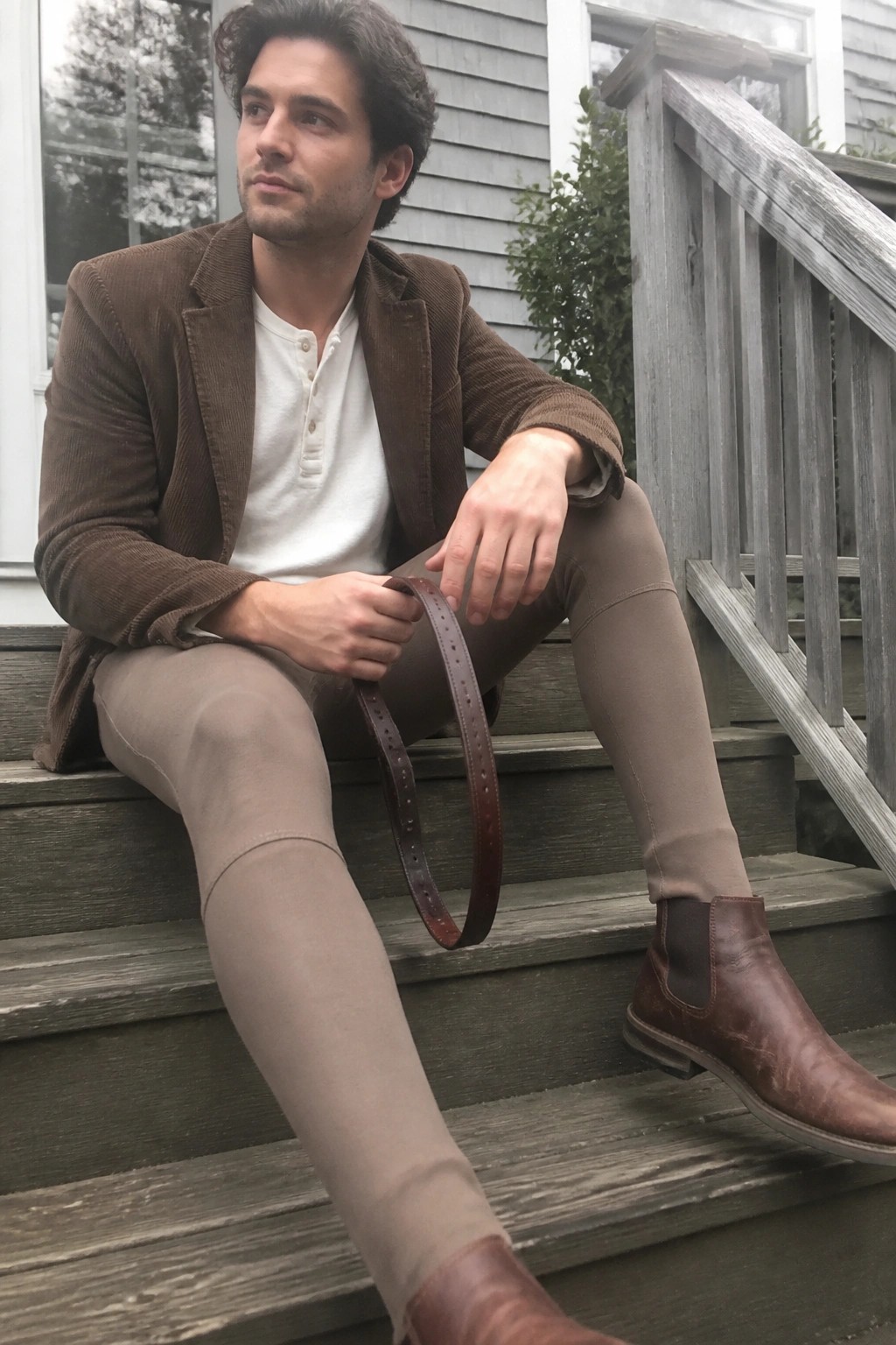 Man in brown corduroy jacket and taupe pants holding leather belt on wooden steps.