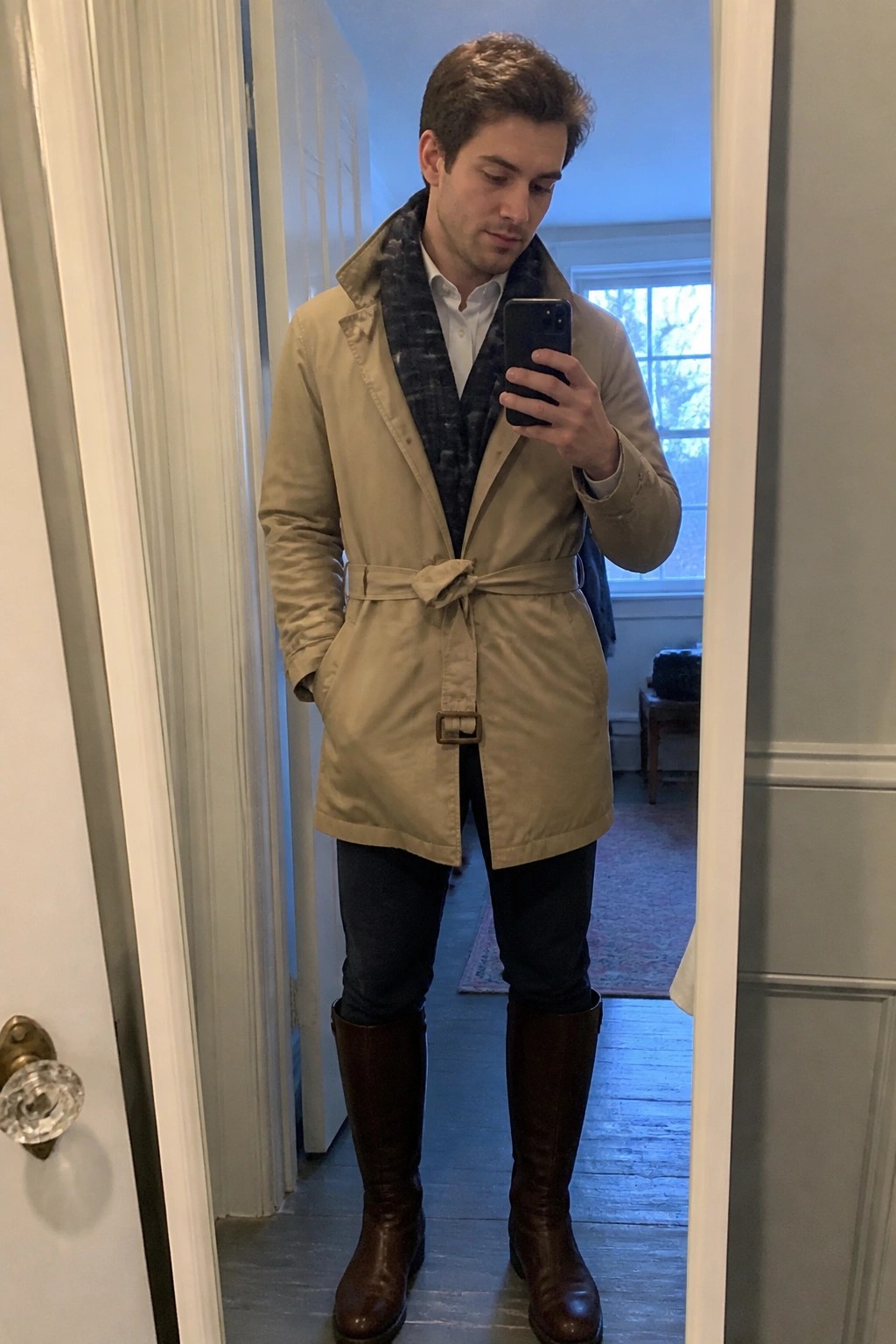Man in beige trench coat with plaid scarf takes mirror selfie wearing brown boots