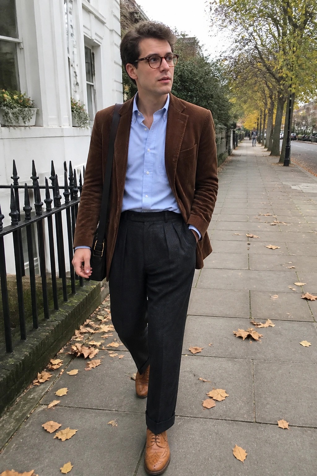 Man in brown corduroy blazer over light blue button down shirt, dark pleated trousers, and brown brogue shoes with black crossbody bag on city sidewalk.