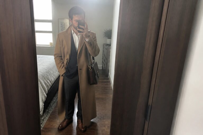 Man in tan overcoat over black vest taking mirror selfie with leather bag