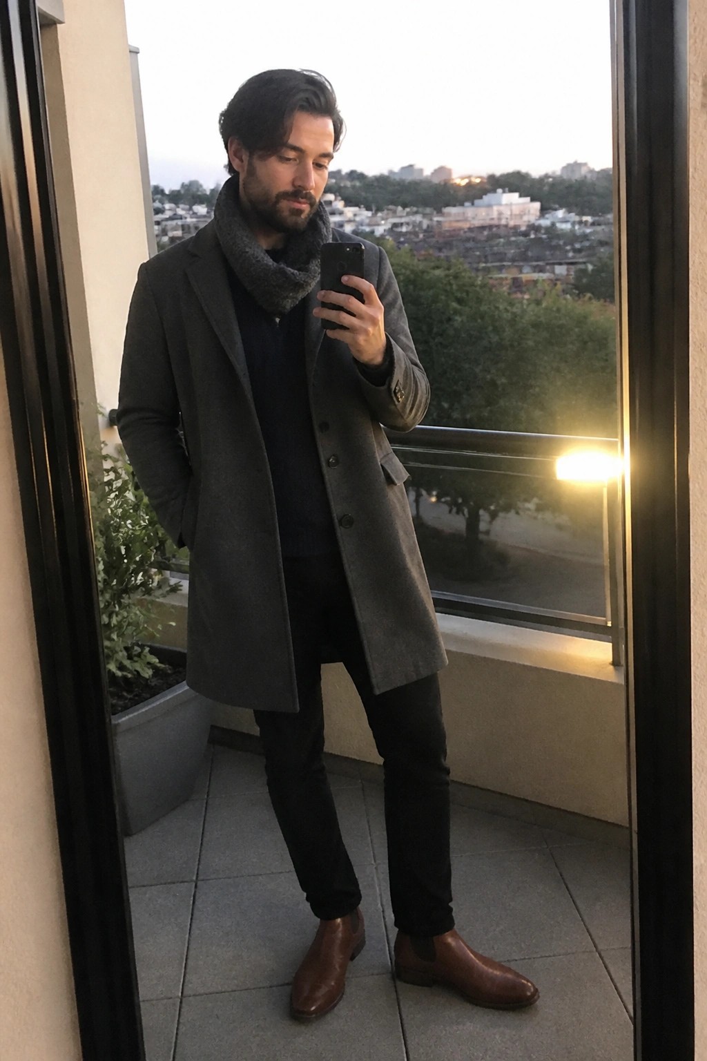 Man in gray coat and scarf taking selfie on dusk city balcony