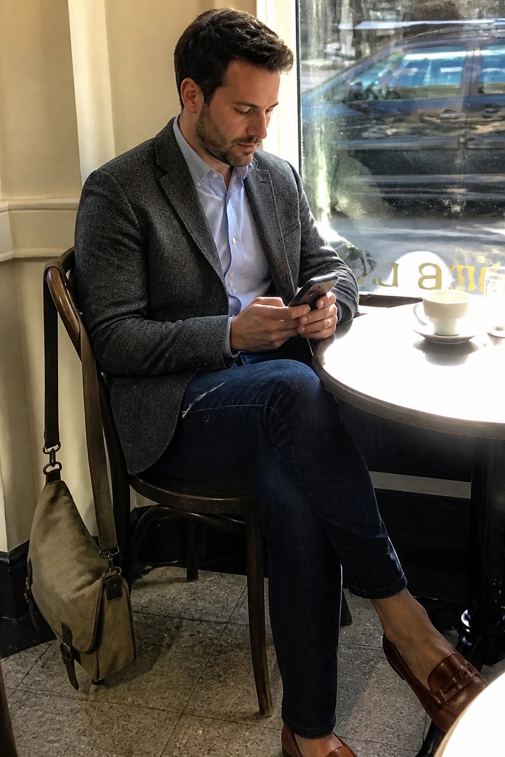 Man seated at a cafe table wearing a gray herringbone blazer over a light blue button down shirt with dark blue jeans and brown loafers while holding a phone.