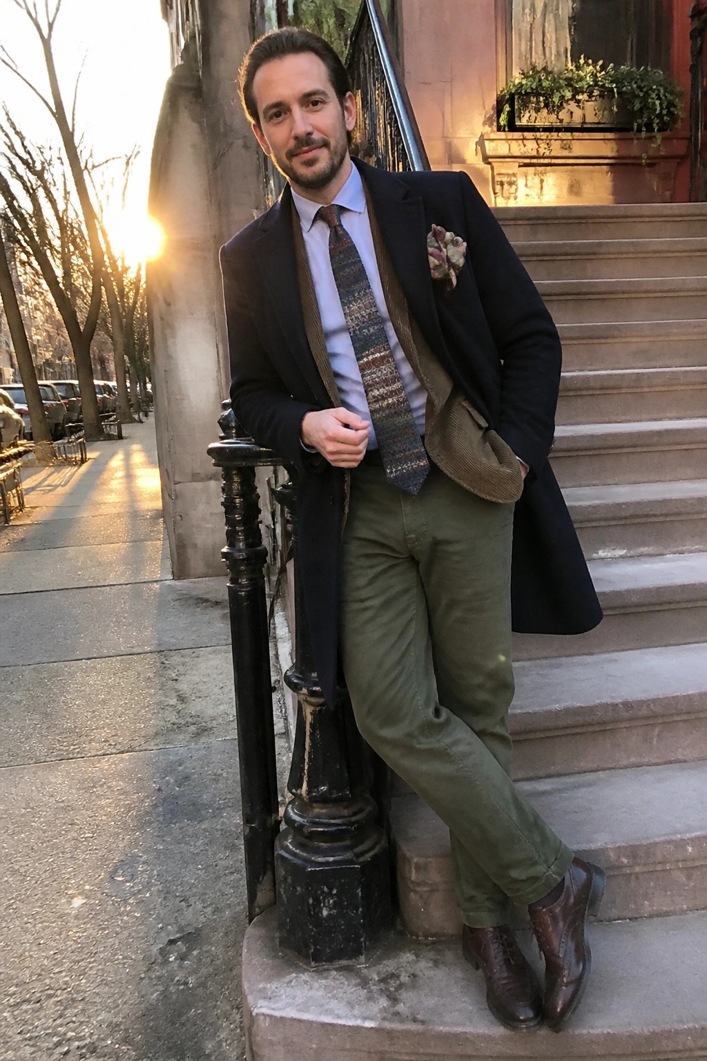 Man in navy blazer, patterned tie, olive pants on urban steps at sunset.