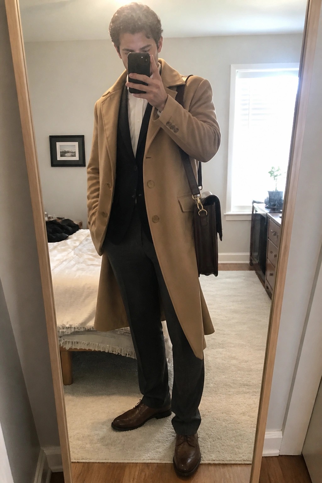 Man in tan overcoat over black vest taking mirror selfie with leather bag