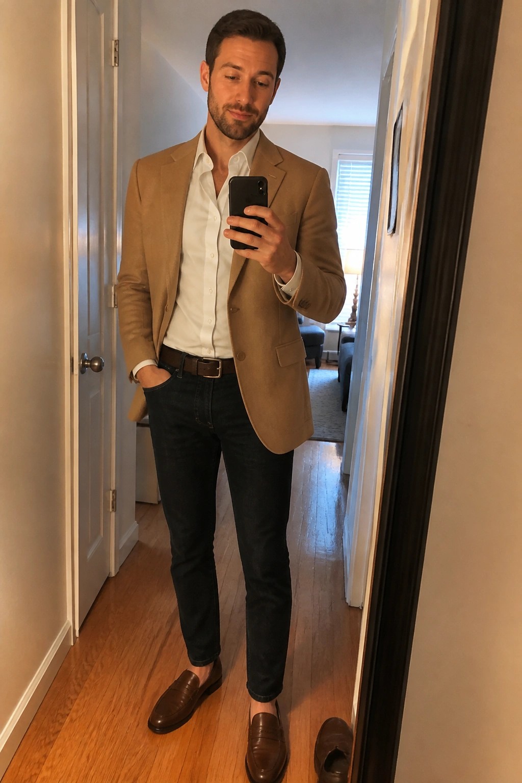 Man in tan blazer, white shirt, and jeans taking mirror selfie in hallway
