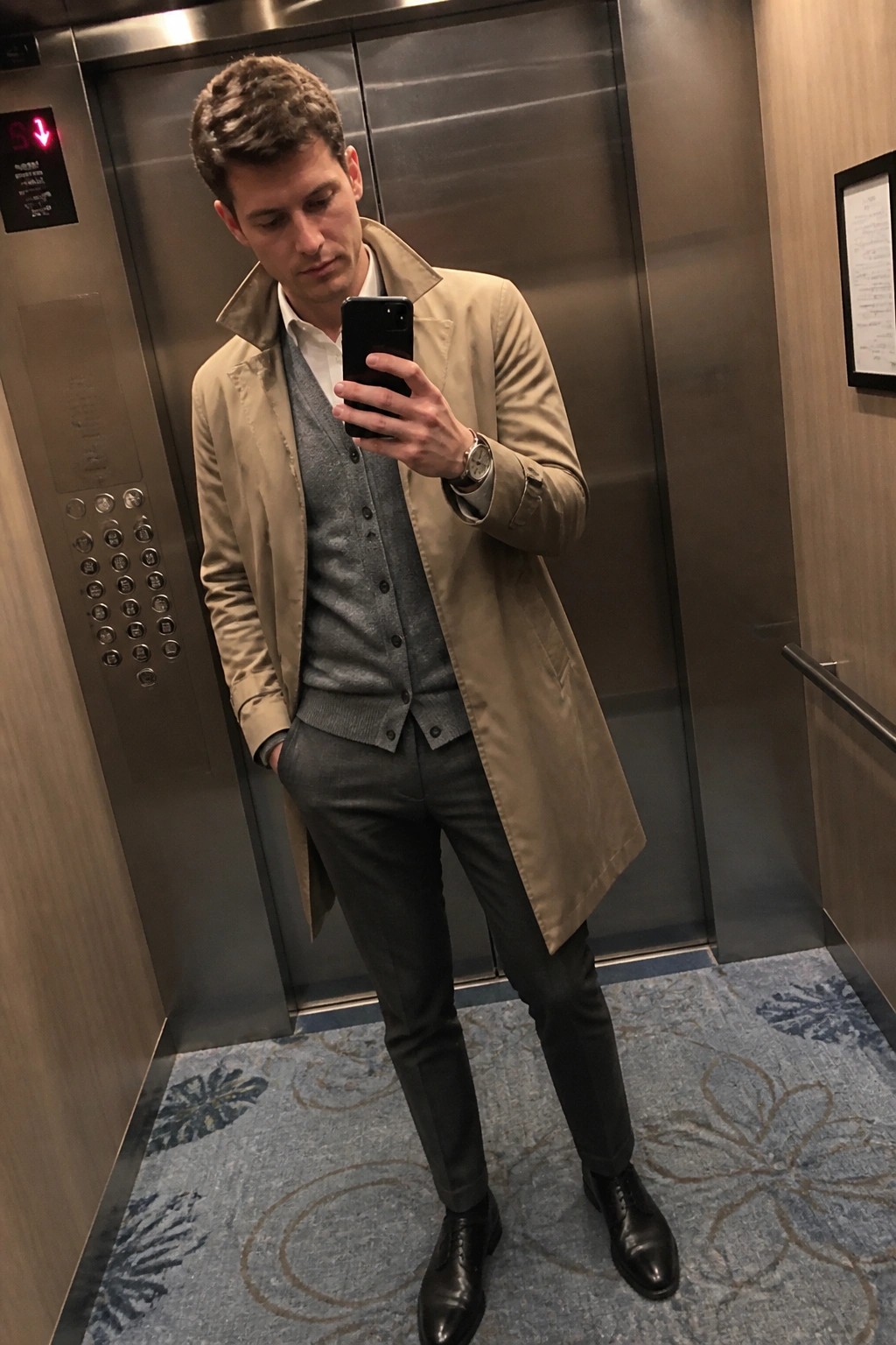 Man in tan trench coat and gray vest taking selfie in elevator