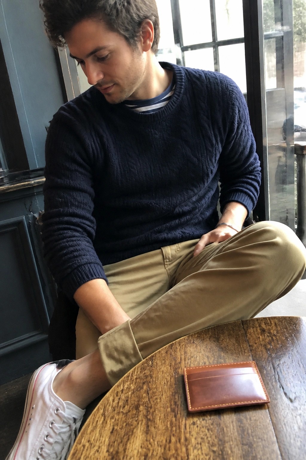 Curly-haired man in navy sweater and khaki pants at table with brown leather wallet.