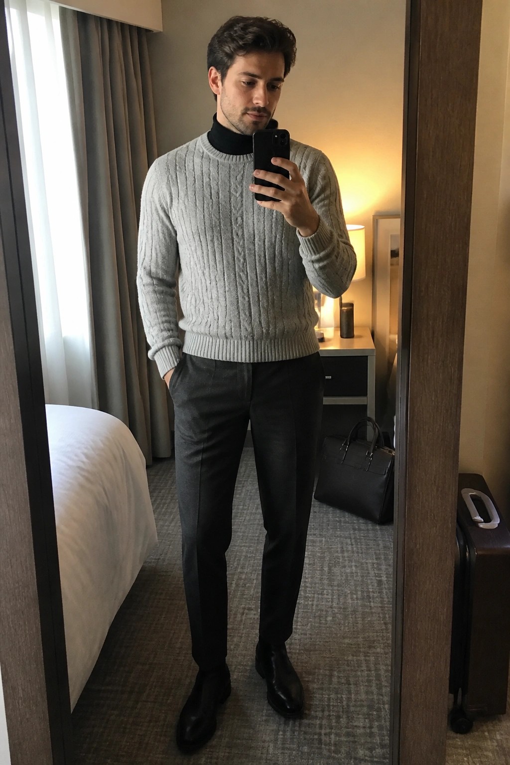 Man in gray cable knit sweater over black turtleneck with black trousers and boots taking a mirror selfie in a hotel room.