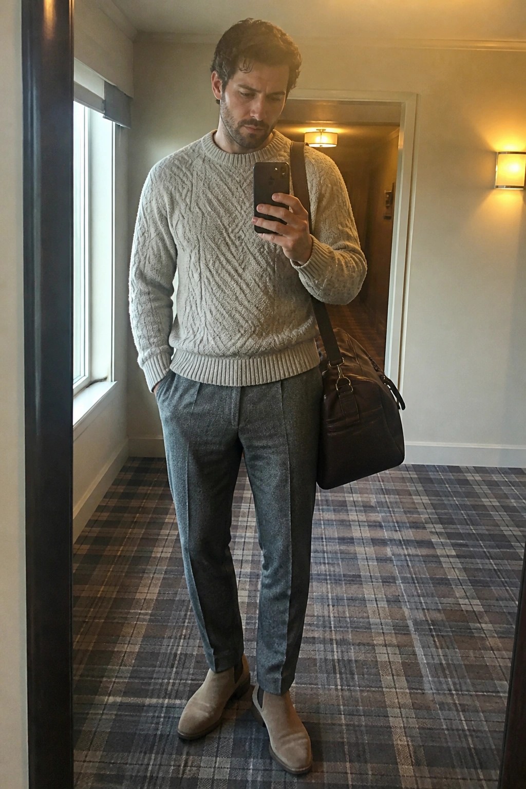 Man in gray cable-knit sweater and trousers taking mirror selfie with leather bag