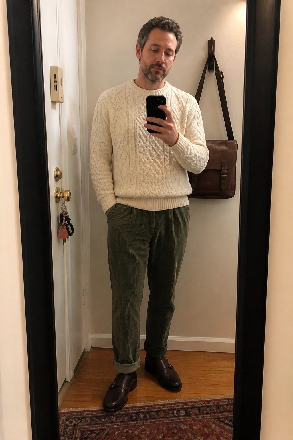 Man in cream cable-knit sweater and green corduroy pants taking mirror selfie.
